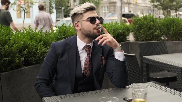 Serious Businessman Smoking Cigar And Drinking Scotch Or Whiskey In Restaurant Terrace, Elite Mens Club Outdoor. Relaxing Time. Man Crushes Cigar Ash