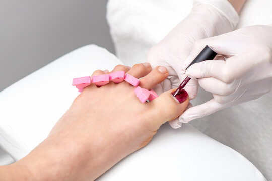 Hands in protective gloves painting red nail polish on female toenail in beauty salon