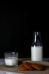 cookies, a bottle and a glass of milk on canvas napkin on white wooden background against black background