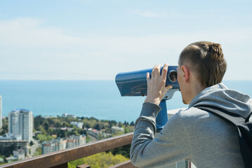 young caucasian hipster man looking through stationary binoculars at sea and city on observation deck, horizontal outdoors stock photo image