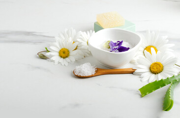 Wooden spoon full of sea salt for bath, bowl with aromatic salt, chamomile flowers, aloe on the white marble table.Empty space