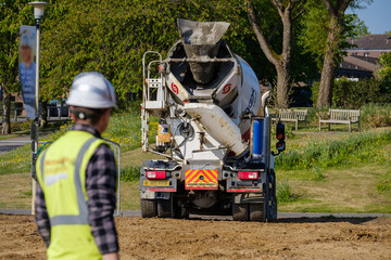 Construction worker in hard hat and hi-vis vest watches as cement truck reverses into construction...