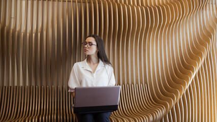 student sits on a wooden bench in the interior and uses a laptop, a young woman