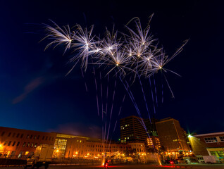 Fireworks. Wide angle shot showing the launch as well as the blooms. Buildings and street lights in the background. Focus is on the fireworks.