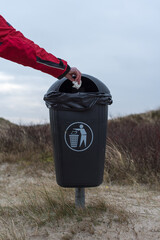 Männerhand wirft ein benutztes Papiertaschentuch in einem schwarzen Mülleimer in den Dünen von Amrum