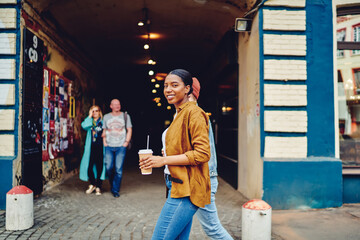 Half length portrait of cheerful African American female tourist with coffee to go in hand exploring city streets and smiling at camera, happy hipster girl with takeaway cup going at urban setting