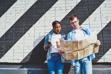 Caucasian young man together with african american hipster girl reading travel map to search right route during trip standing near wall with graffiti.Diverse couple in love tourists using smartphone