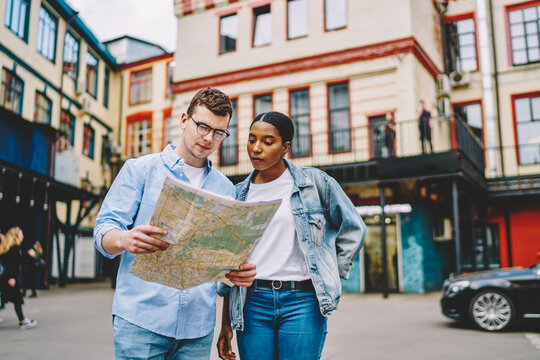 African american young woman together with caucasian friend holding map and searching right direction of route standing on street in urban setting.Trip of multicultural couple in love in new city