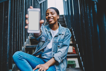 African American millennial female showing smartphone with mockup copy space screen for paste advertising on web content.Cheerful and happy dark skinned blogger holding in hand mobile phone presenting