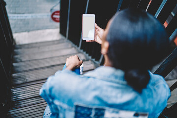 Back view of african american young woman holding modern smartphone with blank screen for your internet content or website sitting on stairs.Female blogger updating app on cellular with mock up area