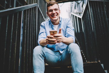 Portrait of cheerful millennial man in classic eyewear for vision protection smiling at camera during mobility networking via application, happy hipster guy in trendy denim apparel holding smartphone