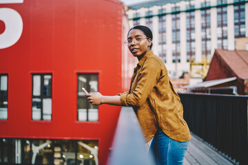 Portrait of millennial female in casual stylish wear spending time on urban setting using smartphone device, dark skinned young woman looking at camera holding cellphone networking via 4G connection