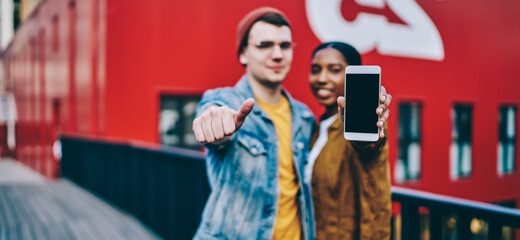 Multiracial hipster guys showing cellphone device with copy space area for internet advertising, blurred millennial male and female promote mock up mobile phone with blank screen for your website
