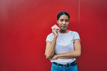 Half length portrait of dark skinned woman in t-shirt with copy space area for brand name or label standing on red wall, thoughtful African American female blogger standing with mobile phone in hand