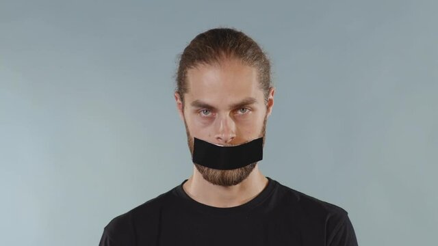Close up man with black tape on mouth over grey background looking at camera serious human shut silence face fear expressive attitude taboo censorship violence scared slow motion