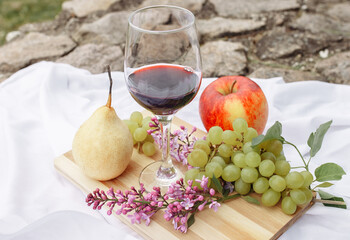 Picnic in the nature in the park. A glass of wine green grapes yellow pear red apple lilac on a wooden board on a white tablecloth in the grass in the garden on a stone path.