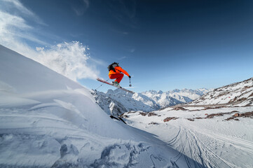 Naklejka na ściany i meble Athlete male skier jumps from a snow-covered slope against the backdrop of a mountain landscape of snow-covered mountains on a sunny day. The concept of winter sports wide angle