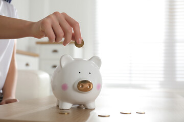 Woman putting money into piggy bank at wooden table indoors, closeup. Space for text