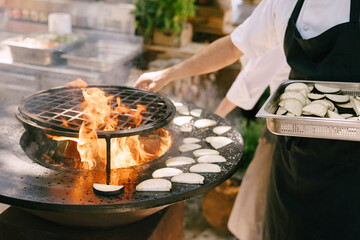 The cook roasts the courgettes. Grilled vegetables. Round bowl-shaped grill with a fire inside. 