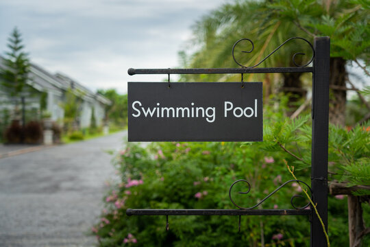 "Swimming pool" direction sign on black metal plate with background of luxury house village and greenery garden environment. Close-up sign/symbol object photo.