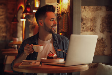 Young blogger with laptop eating dessert and drinking coffee at table in cafe