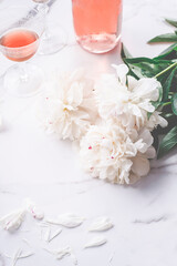 Rose wine in glasses and beautiful white peonies and a scattering of petals on a white marble background. Romantic dinner concept