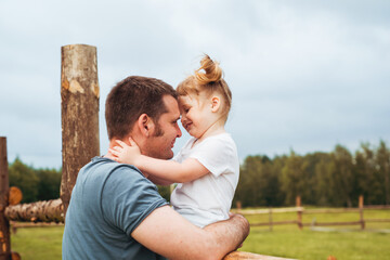 Obraz premium Father andbeautiful baby girl in the village. Sitting high on a wooden fence. Daughter hugs and kisses her father. tender happy family. father's day.