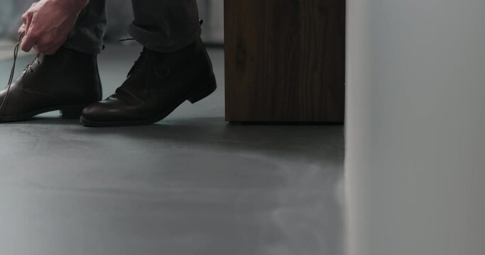 Slow motion dolly shot of young man lace up his brown leather chukka boots indoor