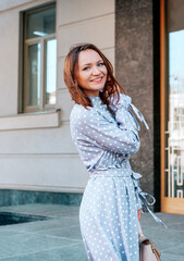  stylish young beautiful woman with red hair in a gray dress is smiling on a city street