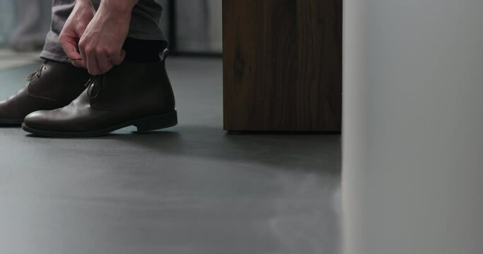 Slow motion dolly shot of young man unlace his brown leather chukka boots indoor