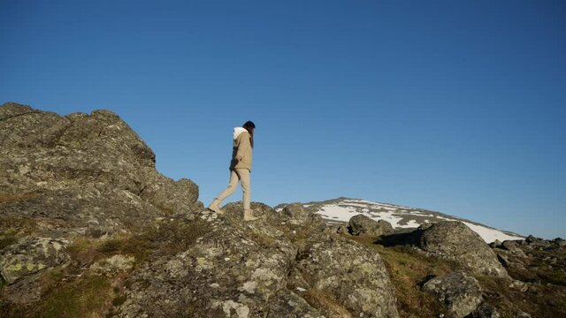 A brunette girl in a beige suit wanders among a large number of stones in the mountains under the sun. Tourist among the mountains. Walk to the top of the mountain.