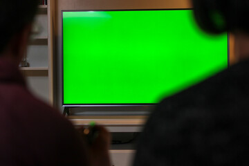 Woman and man are sitting next to each other at home looking at Green Chroma Key Screen, Relaxing on a Couch. Husband holds remote controller in his hand. Couple is watching TV Concept.