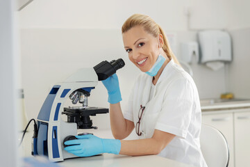 Fototapeta na wymiar Side view of dedicated blond female lab assistant sitting in laboratory and using microscope for blood test.