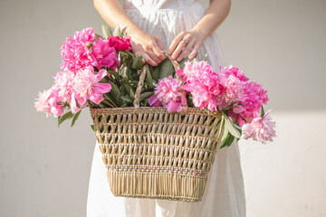 Boho girl holding pink peonies in straw basket. Stylish hipster woman in bohemian floral dress...
