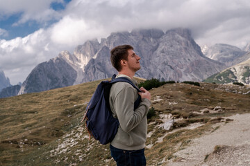 Fototapeta premium Dolomites Alps. Tre Cime di Lavaredo. Italy. Brunnete hiker with blue backpack enjoys alpine trail on background of rocky mountain peaks wrapped by grey clouds in summer