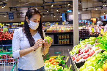 Asian woman wearing protective face mask push and hold shopping cart in supermarket department store. Girl, looking grocery to buy  some fruit. New normal after covid-19. Family concept.
