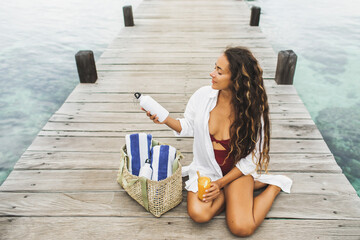 Woman sitting on wooden pier, drinking orange juice and holding white metal reusable bottle. Wicker bag with beach towels. Zero waste concept. - 360681221