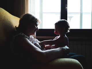 Little girl with pacifier sitting on grandmother's legs in the living room at home - Happy middle-aged grandmother show love and affection - Granny playing relax with granddaughter - Bonding concept