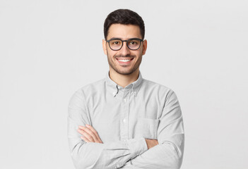 Young business guy wearing gray shirt and eyeglasses, standing with arms crossed, smiling happily...
