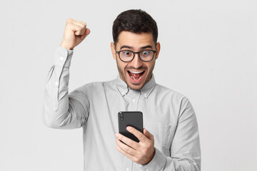 Portrait of excited young business man looking at smart phone, isolated on gray background. Winner...