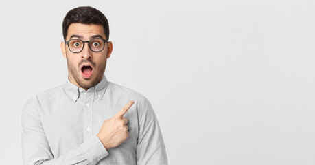 Banner of astonished young man in shirt pointing with finger to right side, isolated on gray background with copy space