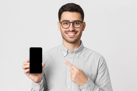 Young man holding mobile phone in hand and pointing to it with finger, isolated on gray background