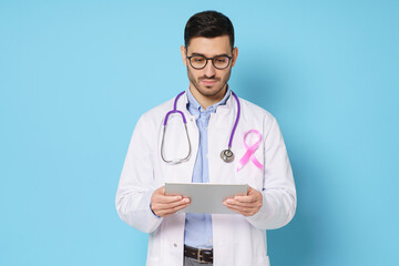 Young male oncologist holding tablet and looking attentively at screen, wearing pink ribbon on his...