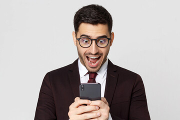 Young business man looking at smart phone with surprise expression, shouting WOW, isolated on gray background