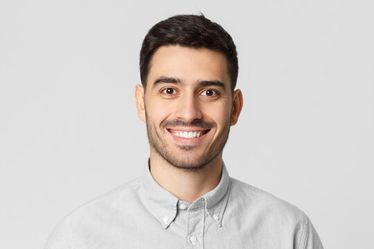 Close up portrait of young smiling handsome man wearing gray shirt, feeling confident as professional, isolated on studio background