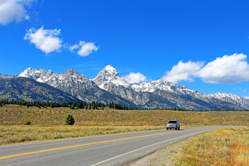 Fototapeta na wymiar Beautiful scenic drive in Grand Teton National park - isolated car driving on the empty road / highway surrounded by high snowy rocky mountain peaks and fields. Wyoming, USA.