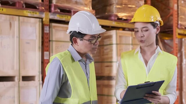asian Male and Female Industrial Engineers in Hard Hats and Safety Jackets Discuss stock inventory while Using data chart. They Work at the stock warehouse Factory