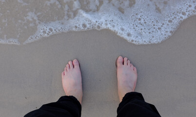 top view man feet in black pants are standing at the edge of the beach with waves of sea water