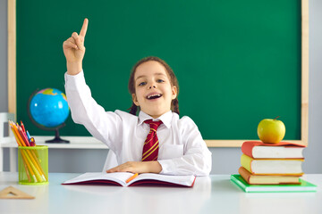 Back to school. Concept idea at school. Cheerful schoolgirl raised her finger up while sitting against a green school board in the classroom.