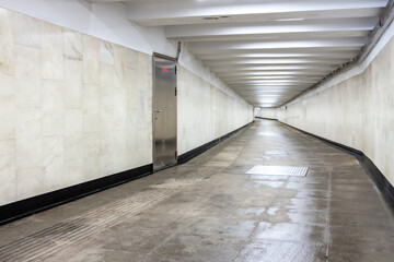 A view down a damp tiled block and concrete tunnel urban or city pedestrian underpass corridor with white wall and lights ceiling.